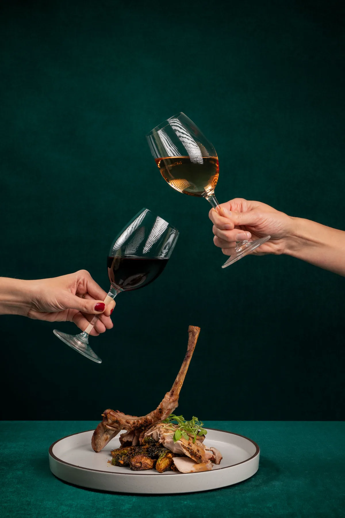 Two wine glasses raised above a plated restaurant dish against a deep green background